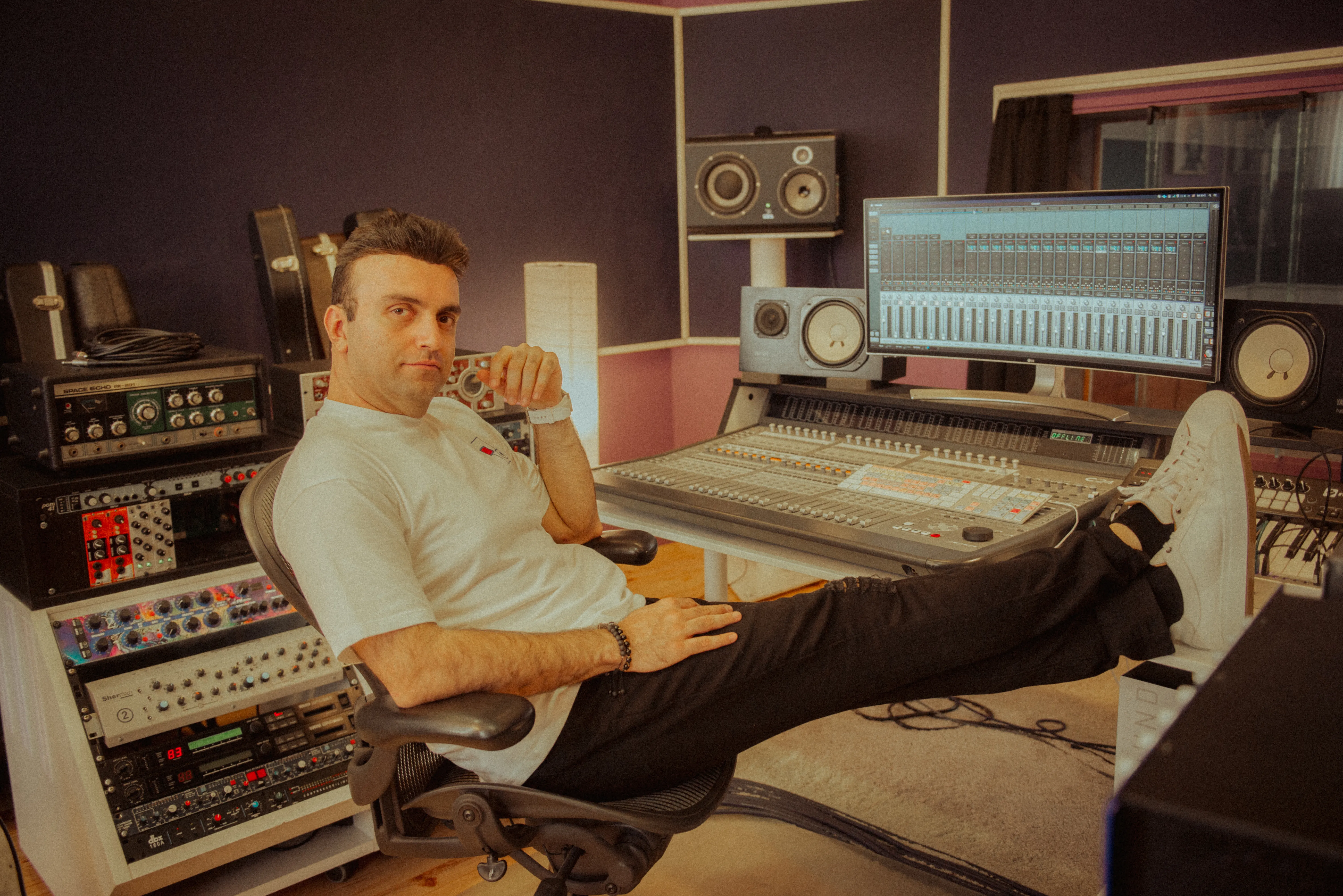 Taha Asadi reclining in his studio chair, feet up beside the mixing console, monitors and rack gear around him