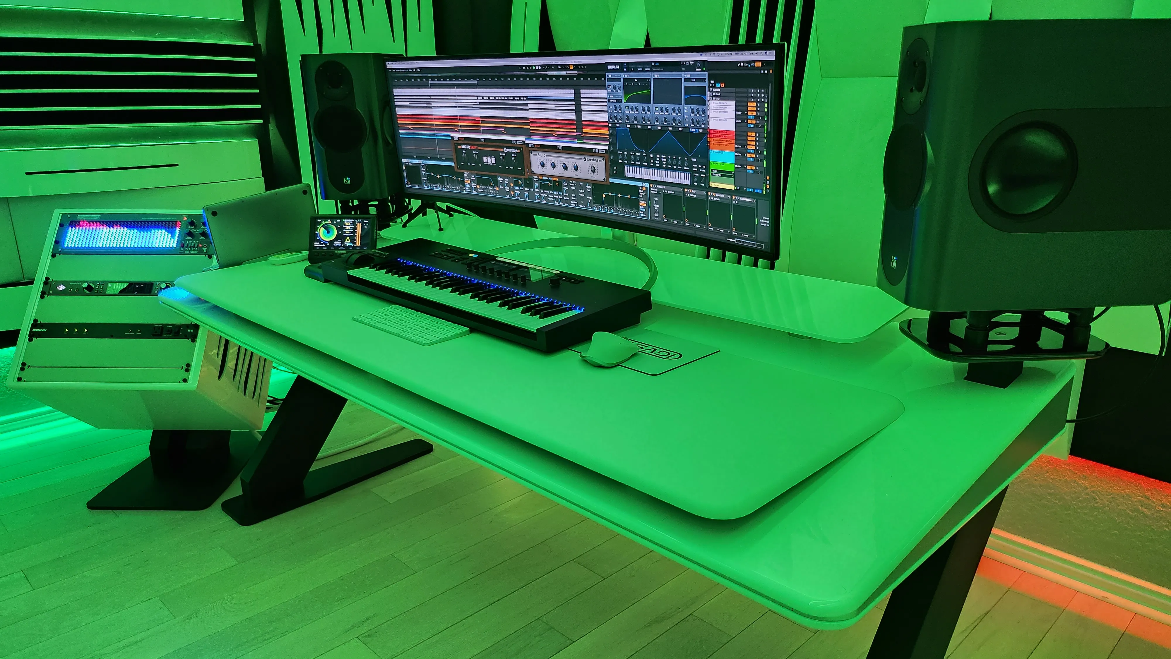 Studio desk washed in green LED light: an ultrawide monitor running a DAW session, a MIDI keyboard, studio monitors on stands, and a sidecar rack beside sculpted acoustic panels