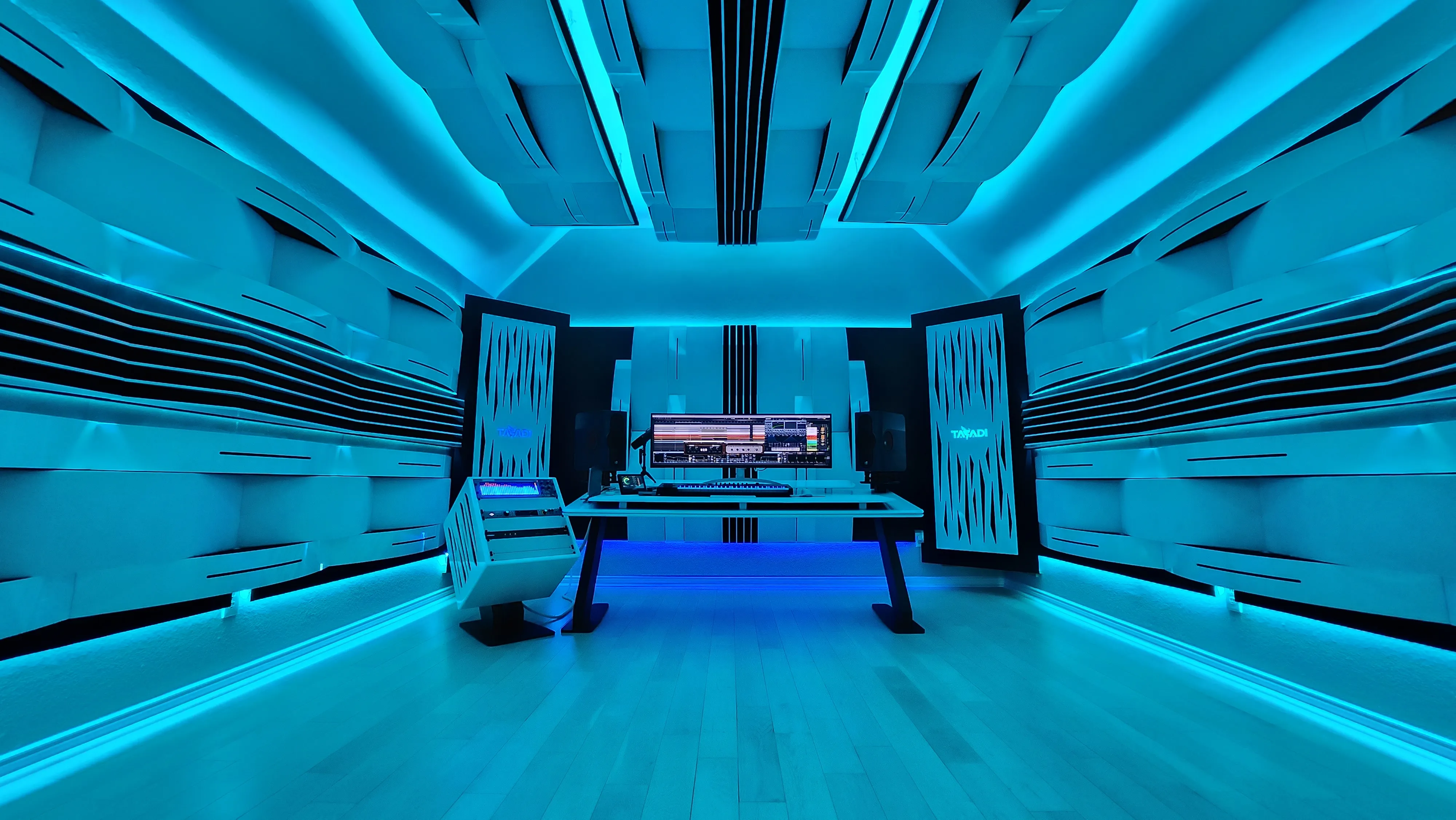Wide view of the studio control room lit in blue and cyan LEDs, acoustic panels lining the walls and ceiling around a centered desk with monitors and a rack unit